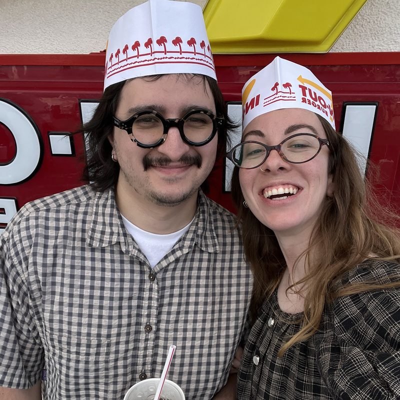 Simón and Tracy in paper hats and matching round glasses, smiling at the camera