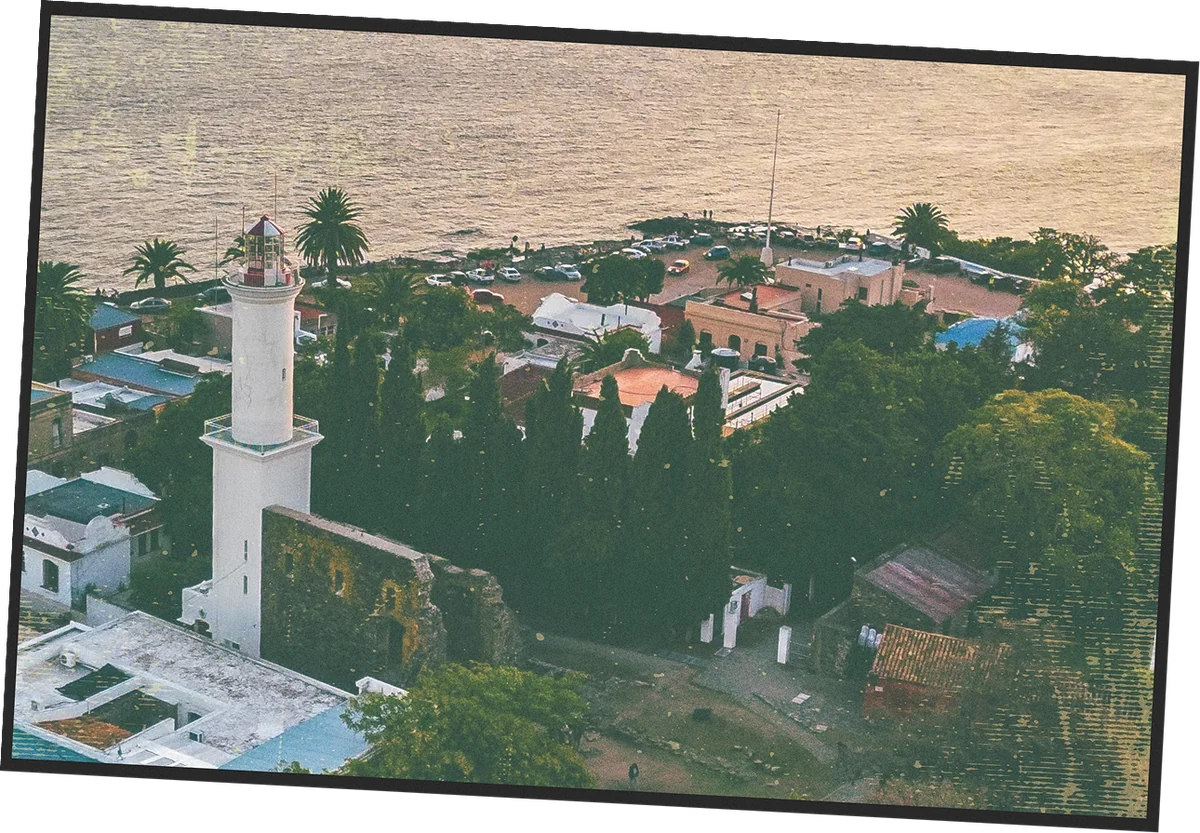 Postcard of a white lighthouse among old rooftops and cypress trees beside the river in Uruguay